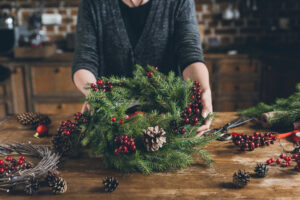 cropped view of florist holding Christmas wreath of fir branches, decorative berries and pine cones at workplace